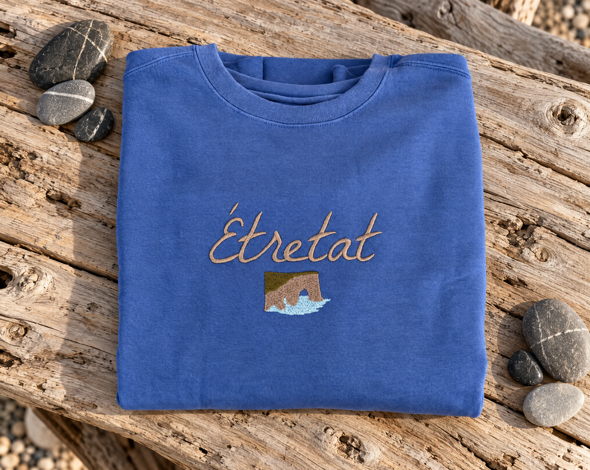 Folded blue Étretat shirt with cliff and wave embroidery on driftwood beside pebbles.