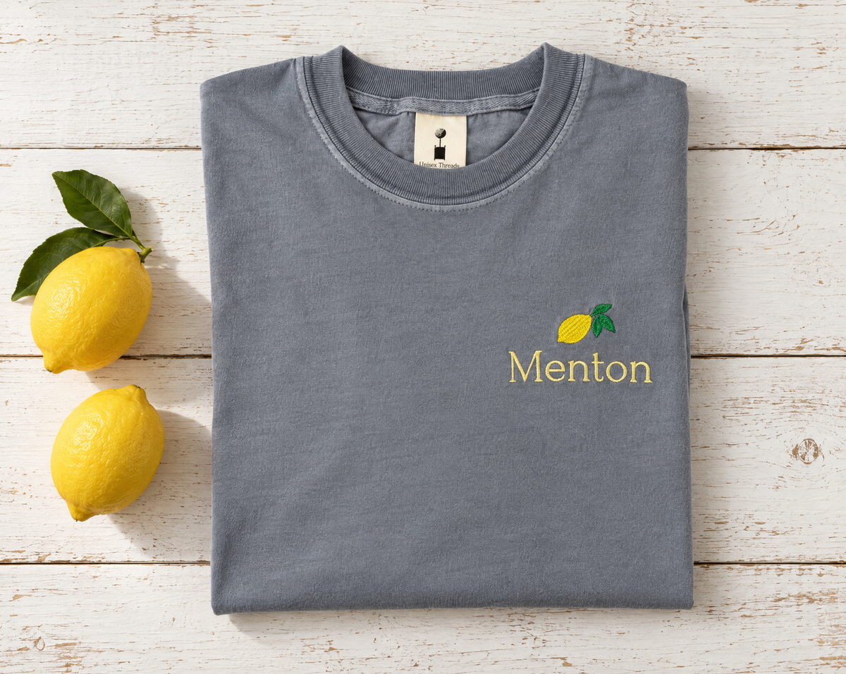 Folded grey Menton shirt with lemon embroidery beside fresh lemons on white wood.