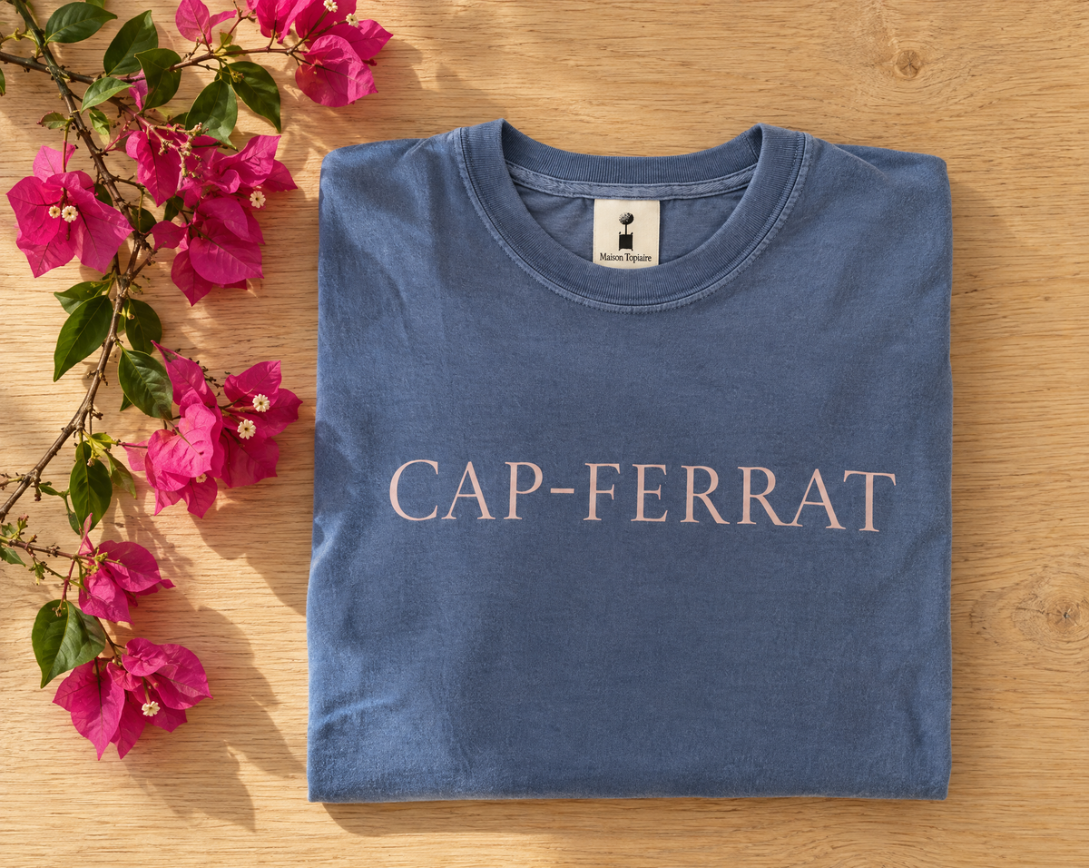 Folded blue Cap-Ferrat shirt with pale pink lettering beside pink bougainvillea on a wooden table.