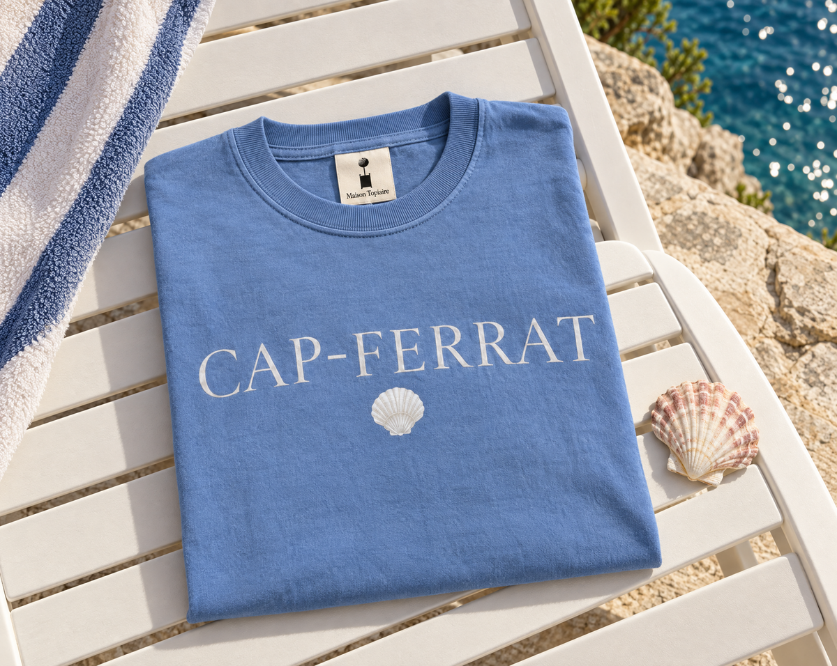 Folded blue Cap-Ferrat shirt with white lettering and a shell print on a white chair beside the sea.