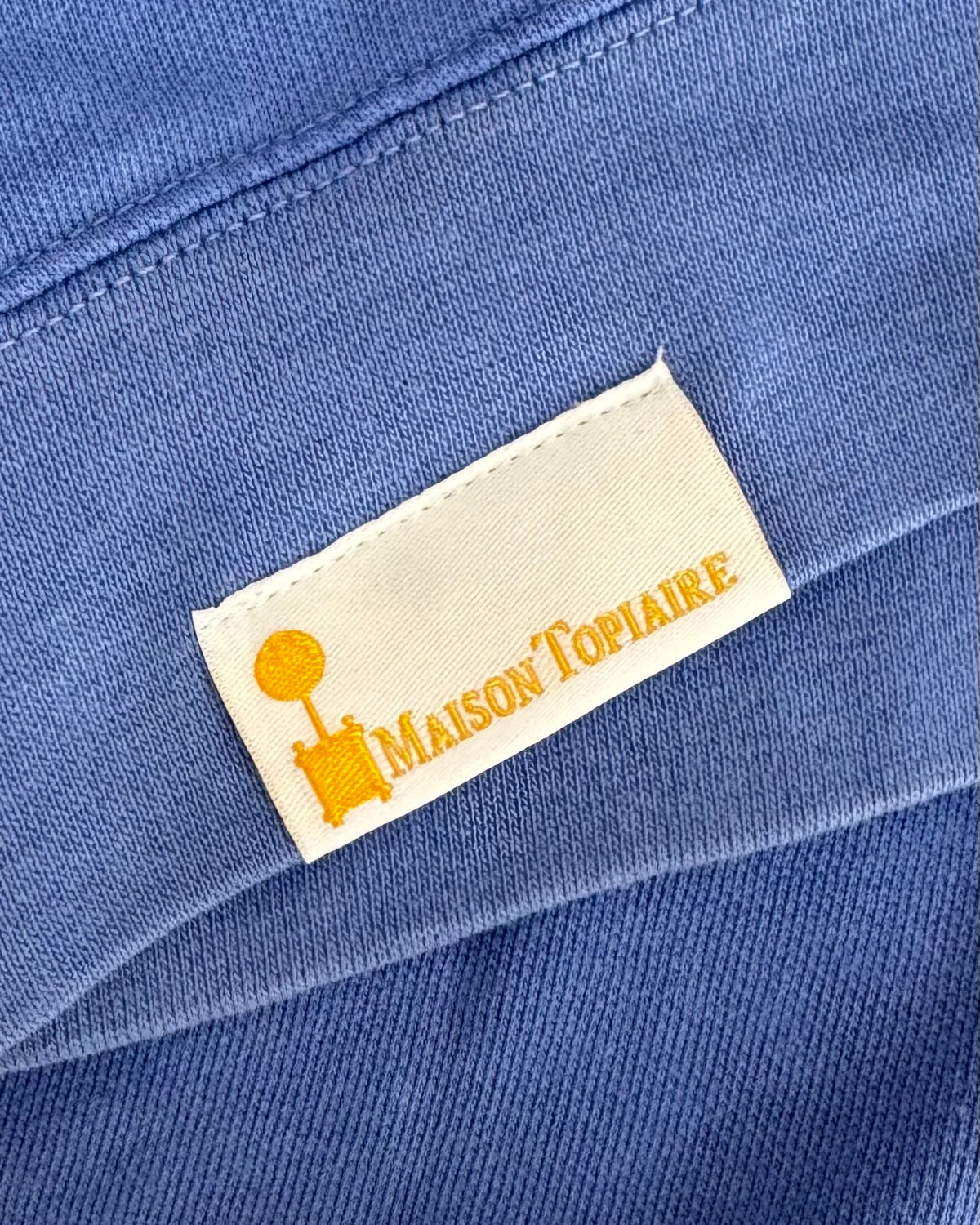 Outward side of the Maison Topiaire woven label shown on a shirt, featuring the topiary logo and brand name